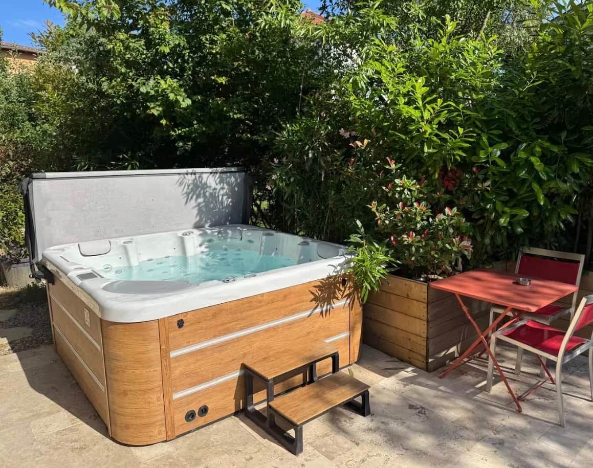 A hot tub with a wooden exterior, a table, and chairs in a garden setting.