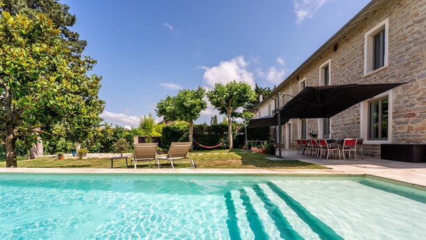A swimming pool with clear water, surrounded by lounge chairs and a stone building with windows.