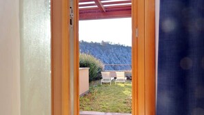 Eine offene Tür führt zu einer Terrasse mit Tisch und Stühlen, einer Holzterrasse und Blick auf einen Hügel.