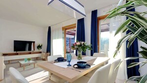 Ein modernes Wohnzimmer mit Esstisch, Stühlen, Fernseher und einem großen Fenster mit Vorhängen.