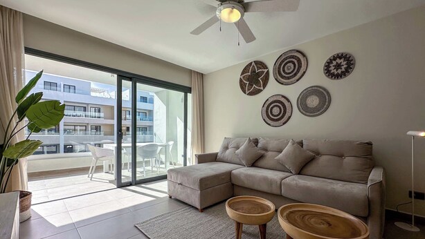Un salon moderne avec un canapé d’angle beige, deux petites tables en bois, une grande fenêtre donnant sur un immeuble et des œuvres d’art décoratives accrochées au mur.