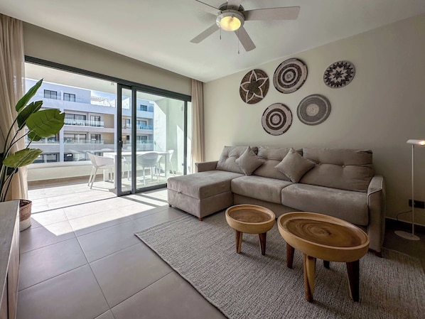 A modern living room with a beige sectional sofa, two wooden side tables, a large window with a view of a building, and decorative wall art.