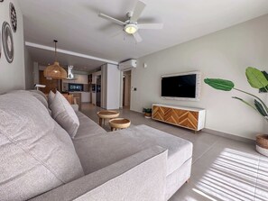 A modern living room with a grey sofa, a TV on a wooden cabinet, a ceiling fan, and a kitchen in the background.