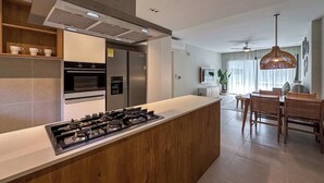 Une cuisine moderne dotée d’un îlot central, d’un four encastré et d’appareils électroménagers en acier inoxydable. On y trouve un coin repas avec une table et des chaises, ainsi qu’un luminaire suspendu.