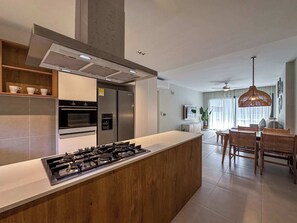 A modern kitchen with a central island, built-in oven, and stainless steel appliances. There is a dining area with a table and chairs, and a hanging light fixture.