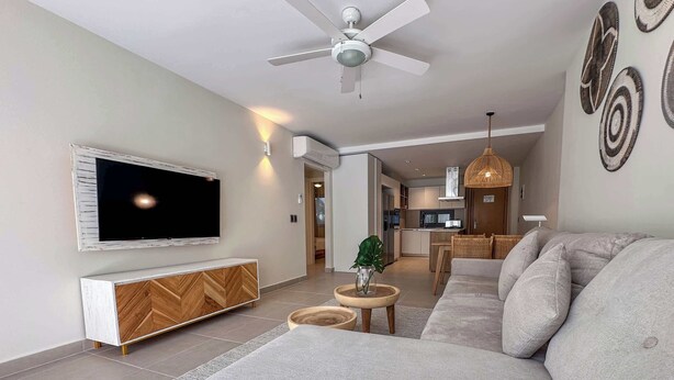 A modern living room with a large sofa, a TV mounted on the wall, a ceiling fan, and a kitchen in the background.