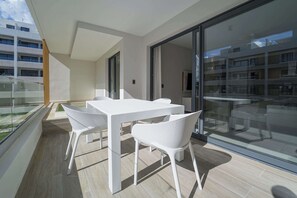 Un balcón con una mesa y sillas blancas, un ventanal de vidrio que da a una habitación y vista a un edificio de la calle.