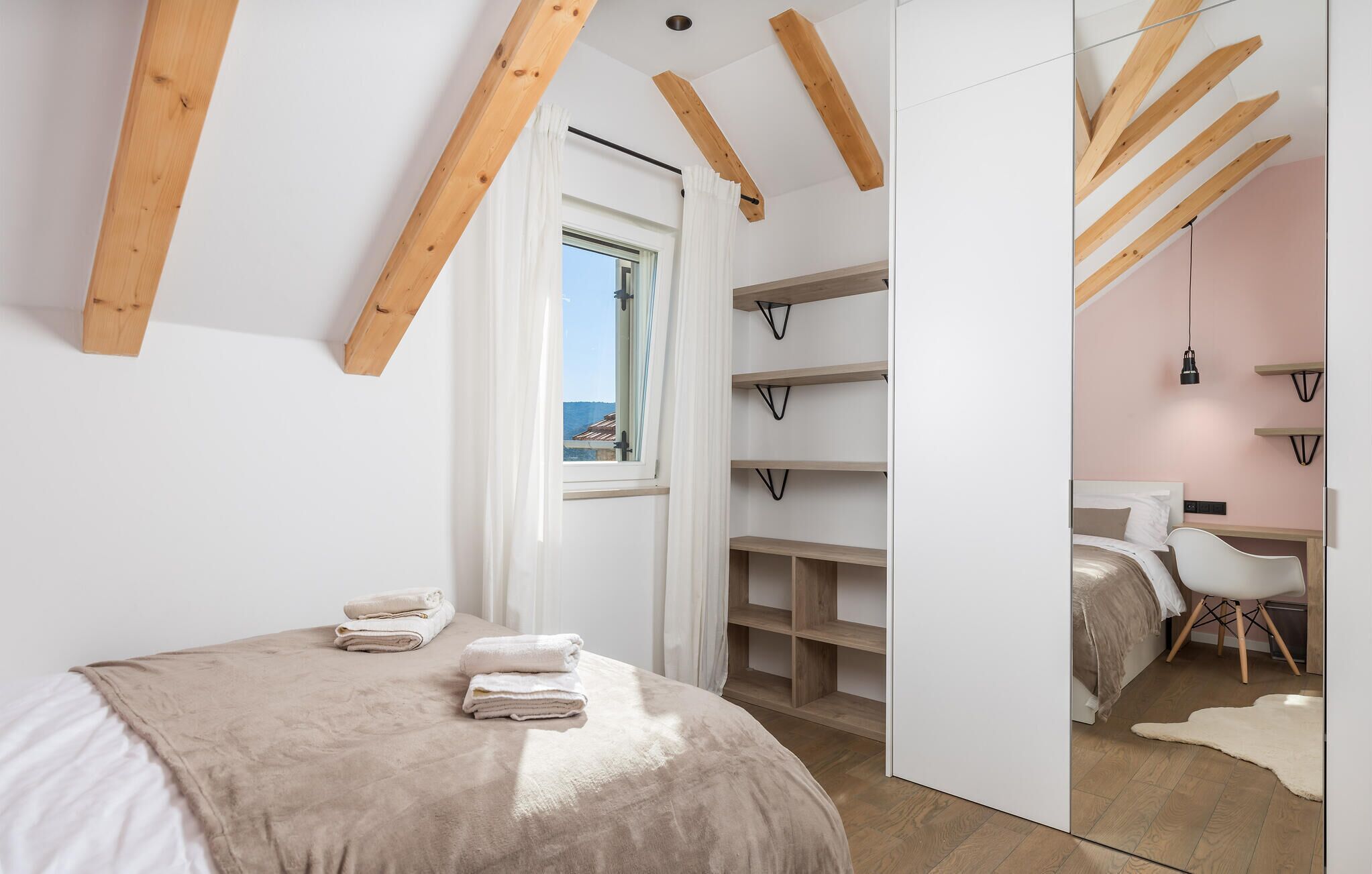 Een slaapkamer met een bed, een bureau en een stoel. Er zijn houten balken in het plafond en een raam met uitzicht op het omliggende landschap.