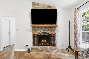 Una sala de estar con chimenea de piedra, un televisor de pantalla plana montado encima y un ventanal con cortinas estampadas.