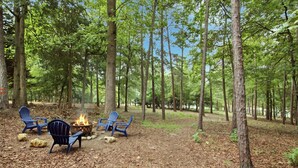 Un campamento en el bosque con un hogar y tres sillas Adirondack.