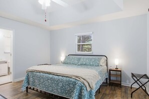 A bedroom with a bed, a bedside table with a lamp, a window with blinds, and a door.