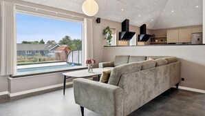 A modern living room with a grey sofa, a dining table, and a large window overlooking a swimming pool and garden. - Stunning home in Spøttrup with sauna (Spøttrup)