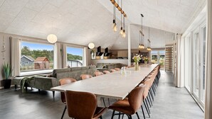 A spacious dining area with a long table and multiple chairs, a kitchen in the background, and large windows offering a view of the outdoors. - Stunning home in Spøttrup with sauna (Spøttrup)