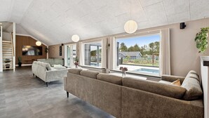 A spacious living room with a large sofa, a flat-screen TV, and a view of a swimming pool. - Stunning home in Spøttrup with sauna (Spøttrup)