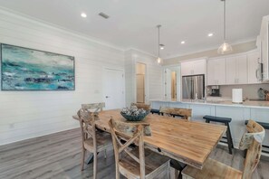 A dining area with a wooden table and chairs, a large painting on the wall, and a kitchen with white cabinets and stainless steel appliances. - Azure Horizon Beachfront Charm in Gulf Shores (Gulf Shores)