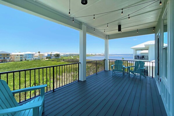 A covered deck with chairs, a railing, and a view of the sea and buildings.