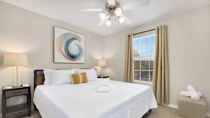 A hotel room with a large bed, two bedside tables, a ceiling fan, a window with curtains, and a painting on the wall.