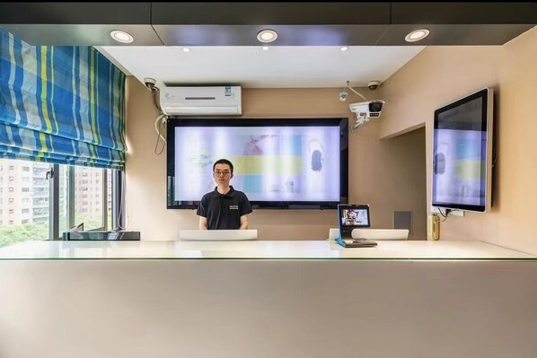 Un área de recepción con una pantalla grande que muestra una imagen desenfocada, un mostrador con una computadora y una persona de pie detrás.