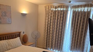 A hotel room with a large bed, a bedside table with a lamp, a wall-mounted fan, and a window with curtains. - 2 Bed Apartment Central London Canary Wharf (London)