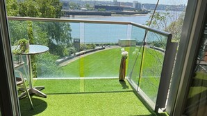 A modern room with a laptop on a wooden desk, a chair, and a view of a river and greenery. - 2 Bed Apartment Central London Canary Wharf (London)