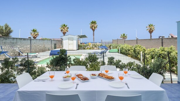 Een terras met buitenmeubilair, op de achtergrond een zwembad, omringd door palmbomen en een heldere lucht.