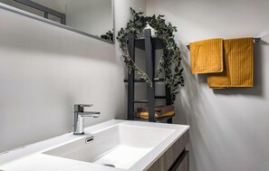 Ein Badezimmer mit einem weißen Waschbecken, einem Spiegel, einem schwarzen Handtuchhalter mit gelben Handtüchern und einer schwarzen Leiter mit Grünpflanzen.