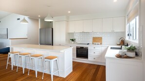 A modern kitchen with white cabinetry, a central island with stools, a stainless steel refrigerator, and a built-in oven. - Seaglass Beach House - Beautifully designed retreat in the heart of Narrawallee (Narrawallee)