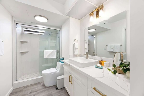 A modern bathroom with a glass shower enclosure, a white toilet, a vanity with a large mirror, and a marble countertop.