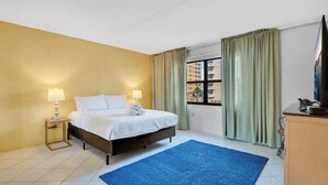 A hotel room with a bed, two bedside lamps, a blue rug, a window with curtains, and a view of buildings outside.