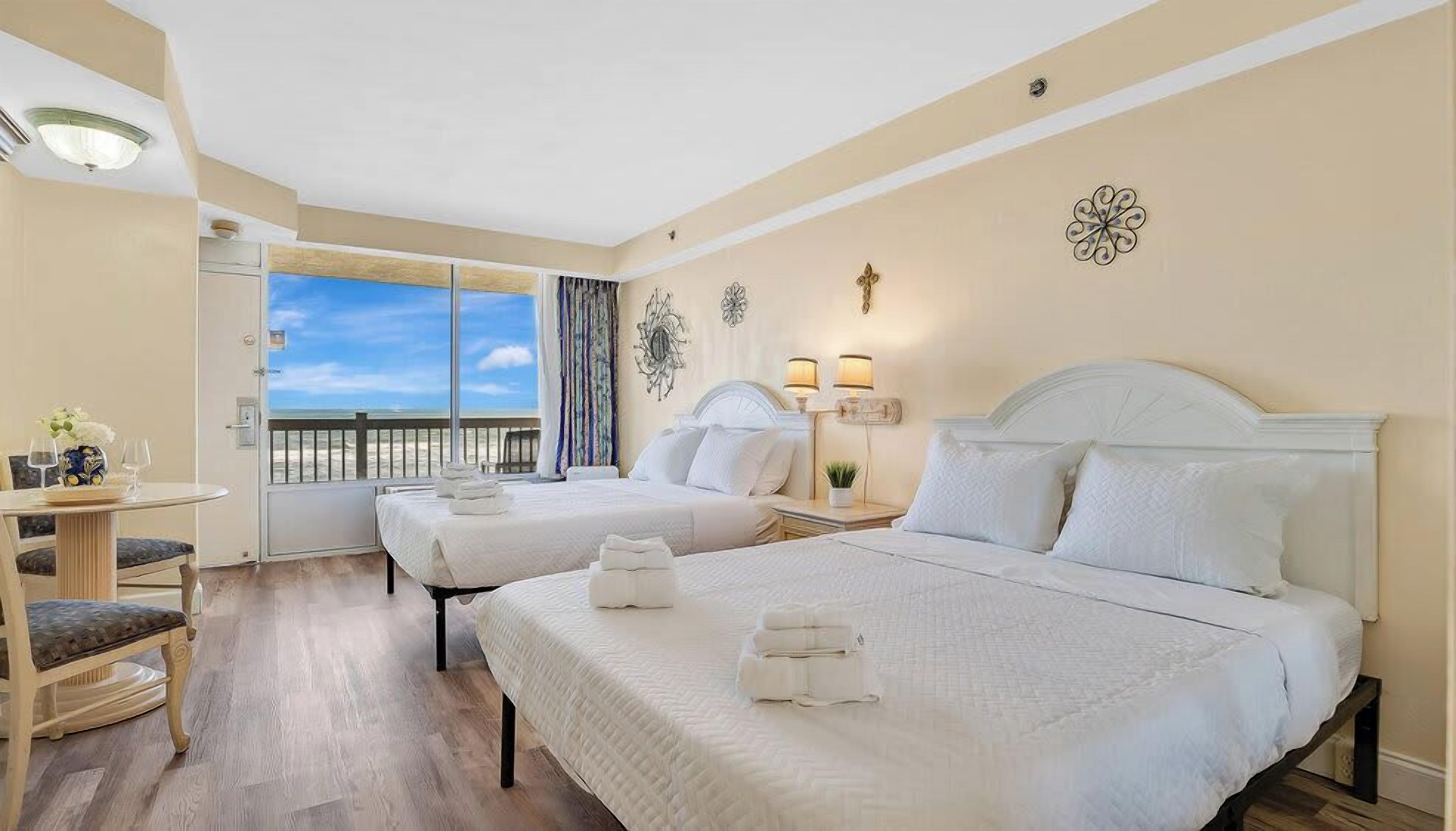 A hotel room with two beds, a small table with chairs, a balcony with a view of the beach, and decorative wall art.