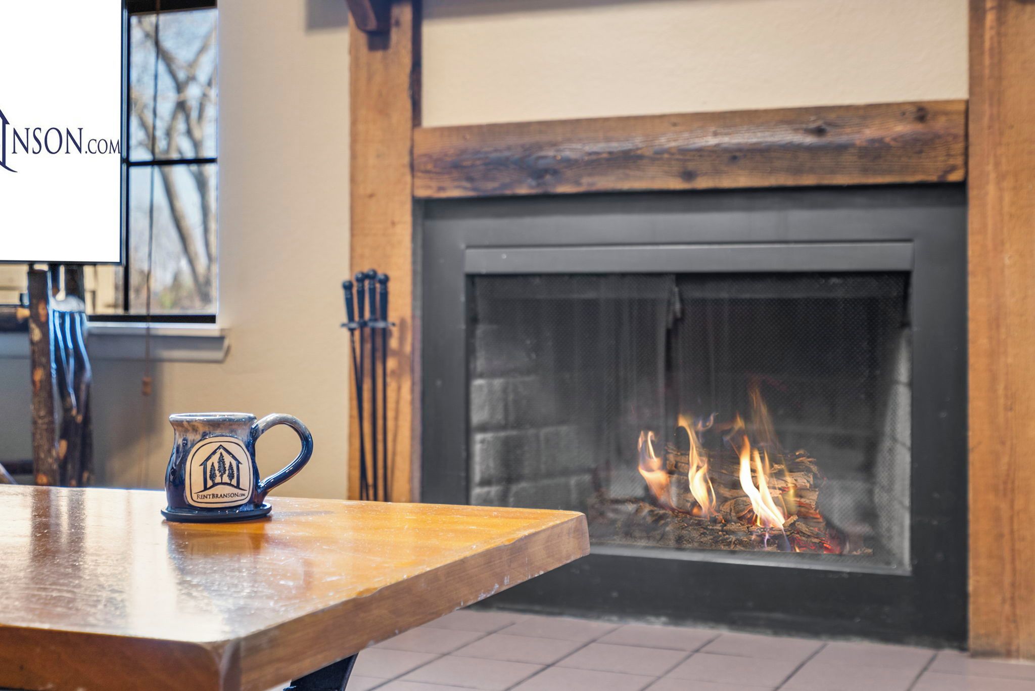 A fireplace with visible flames, a wooden table in the foreground, and a window with a view of trees.