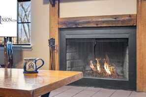 A fireplace with visible flames, a wooden table in the foreground, and a window with a view of trees.