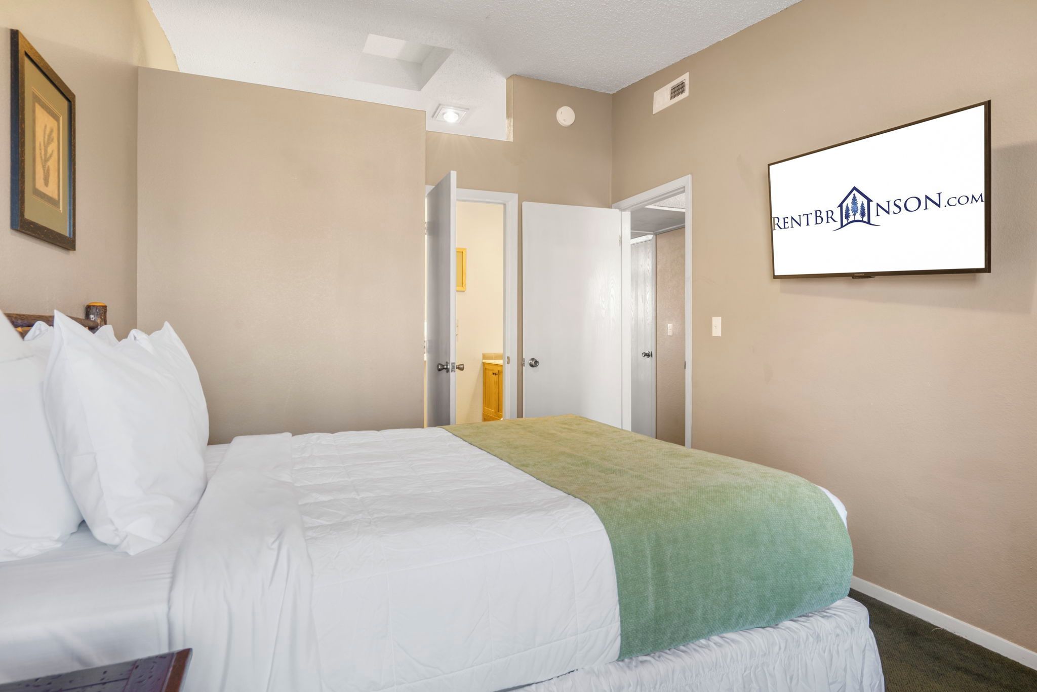 A hotel room with a bed, a green throw, a framed picture, and a sign on the wall.
