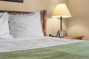 Una cama bien tendida con sábanas blancas, un cabecero de madera, una lámpara de noche, un teléfono y una taza sobre una mesita de noche de madera.