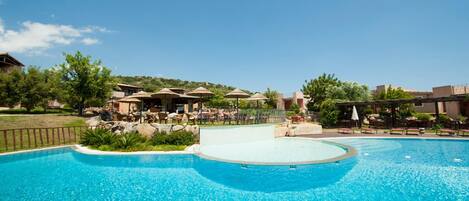 A large outdoor swimming pool with lounge areas and umbrellas, surrounded by greenery and buildings.