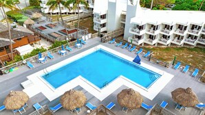 Una piscina grande con tumbonas y sombrillas de paja, rodeada por una terraza de madera y un edificio blanco con balcones.