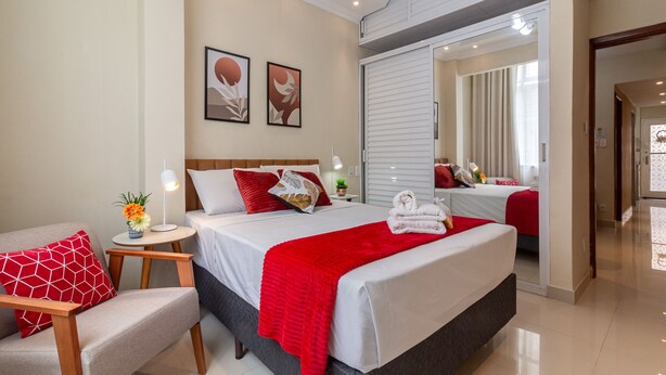 A modern hotel room with a large bed, a seating area with a red and white patterned chair, and a small side table with a lamp and a vase of flowers. - Cosy in Copacabana 7 minutes from the beach | FM211 (Rio de Janeiro)