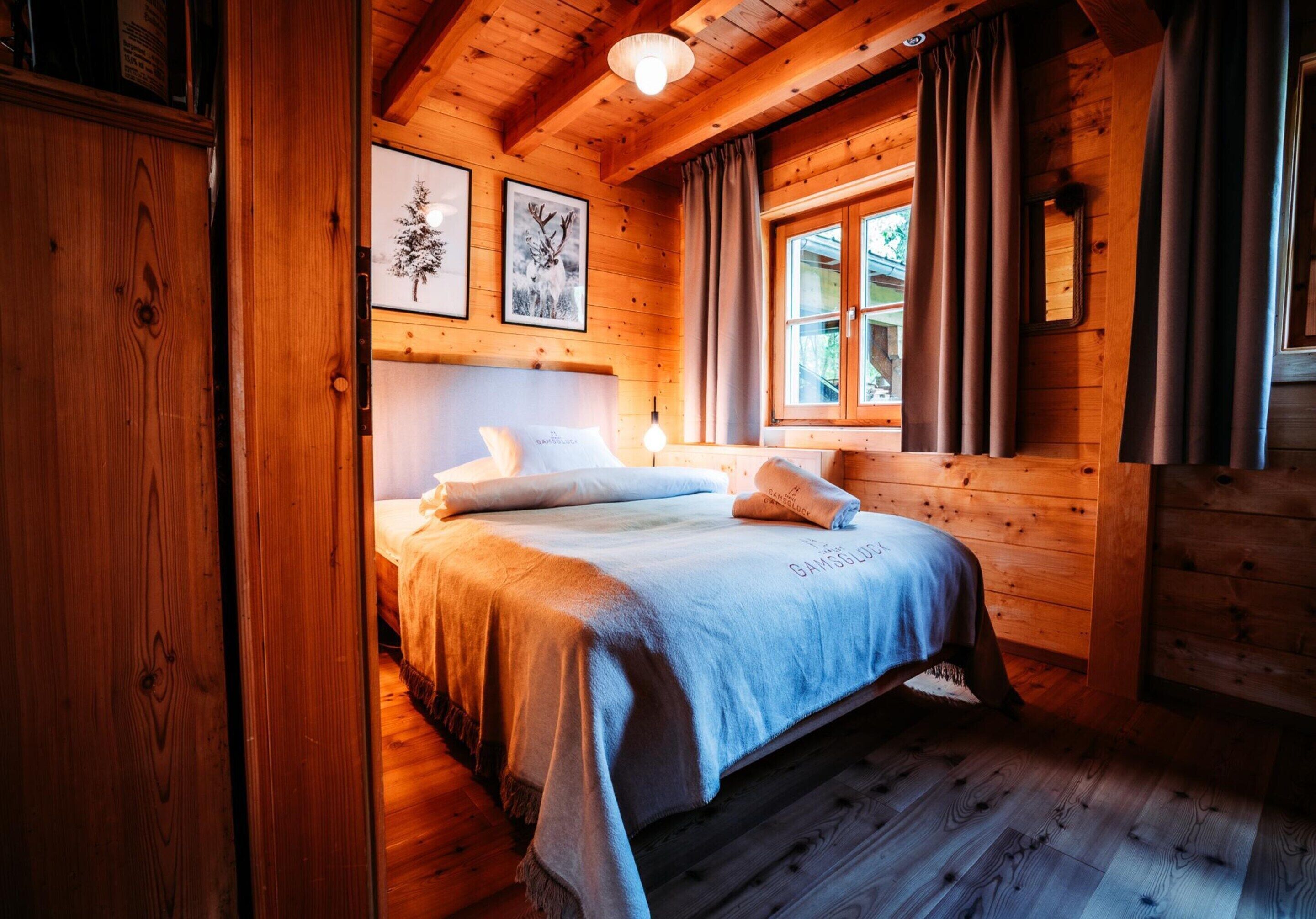 Ein Schlafzimmer einer Holzhütte mit Bett, einem Fenster mit Vorhängen und gerahmten Bildern an der Wand.