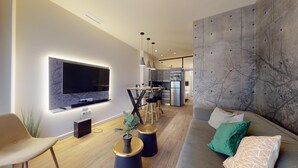 Ein modernes Wohnzimmer mit Flachbildfernseher an der Wand, einem Holztisch, einer Couch mit gemusterten Kissen und einer Küchenzeile im Hintergrund.