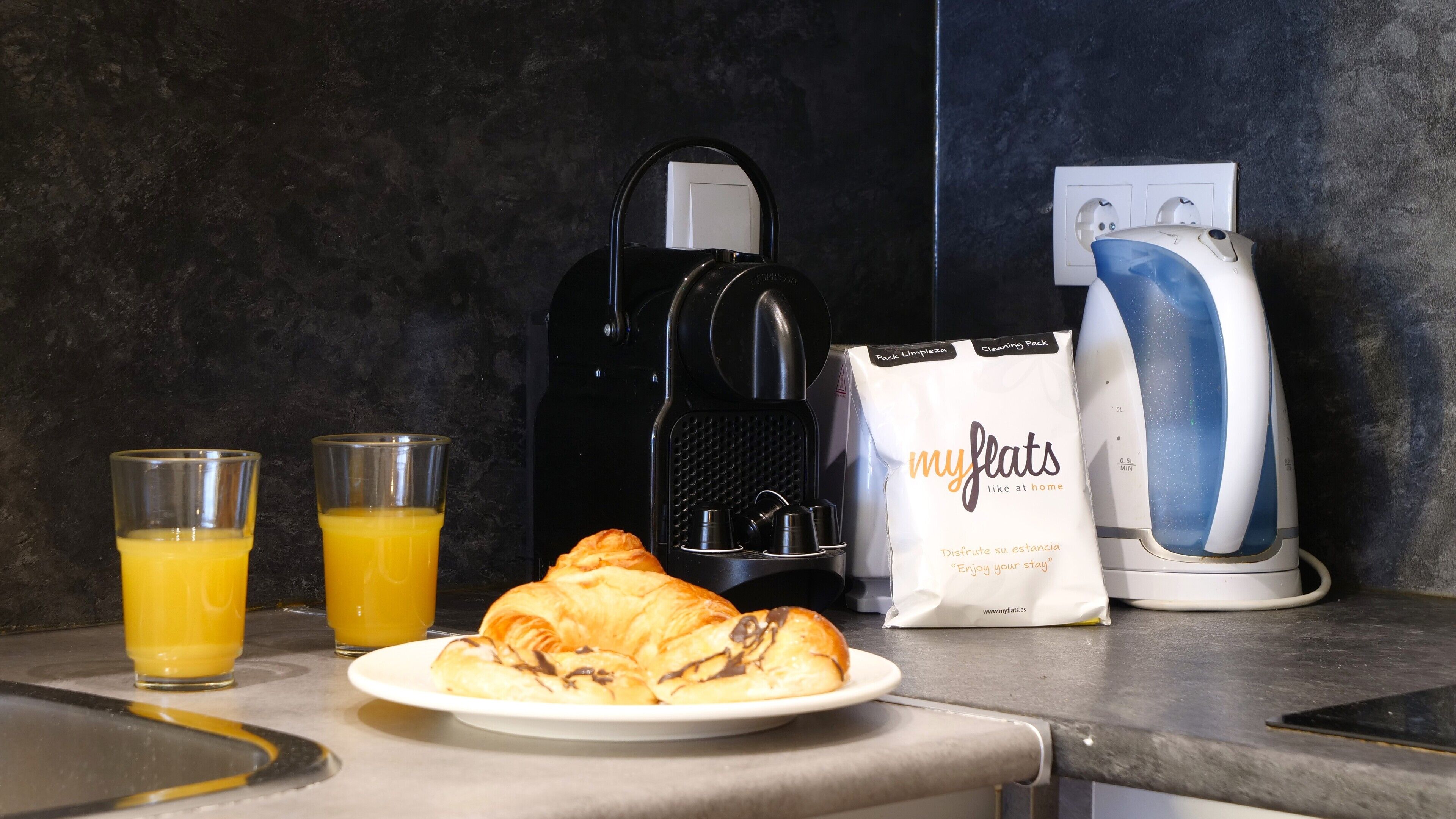 Un bancone della cucina con una macchina del caffè, due bicchieri di succo d'arancia, un piatto di croissant e un kit per la pulizia.