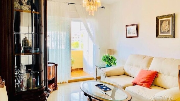 Sala de estar com um sofá bege, uma mesa de centro de vidro, um lustre e um aparador de vidro com objetos decorativos.