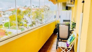 Uma varanda com paredes amarelas, uma cadeira, uma mesinha e vista para a cidade.