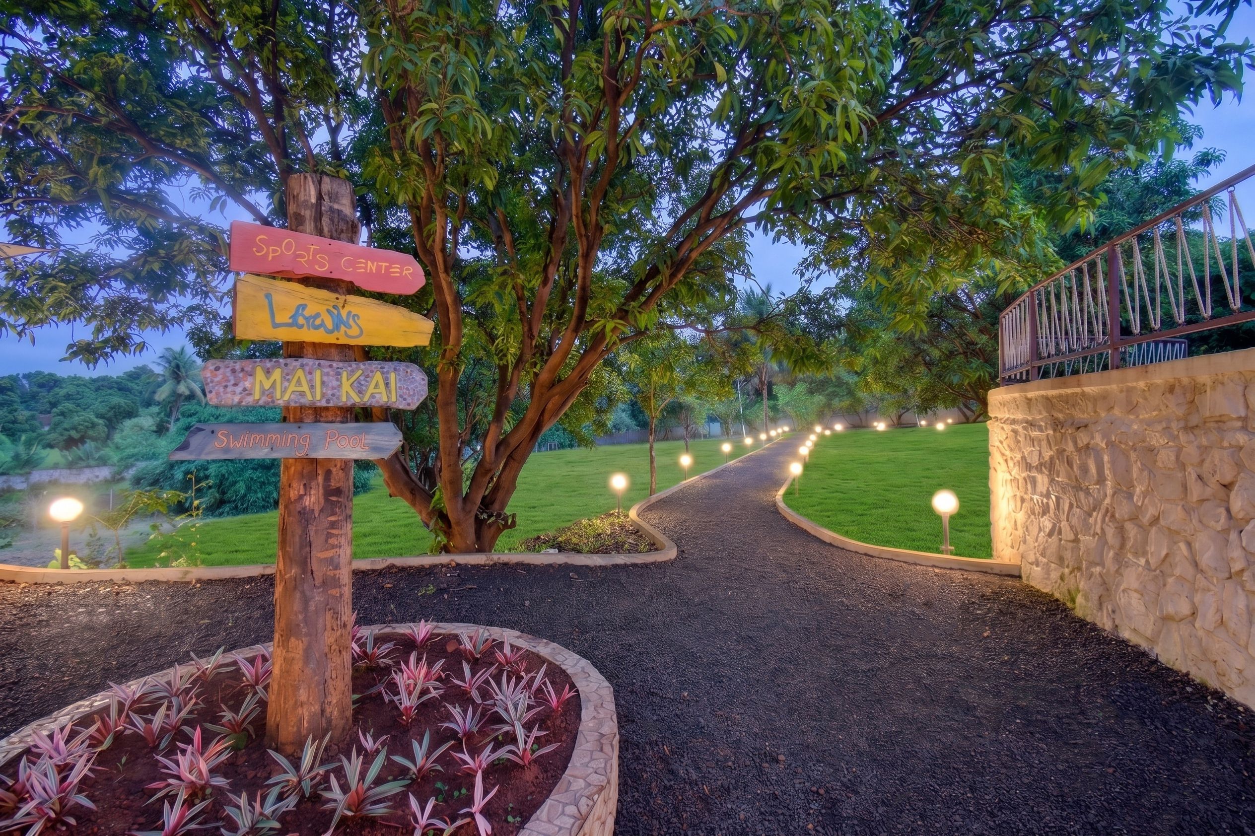 A pathway leading to a sports center and swimming pool, surrounded by lush greenery and a stone wall.