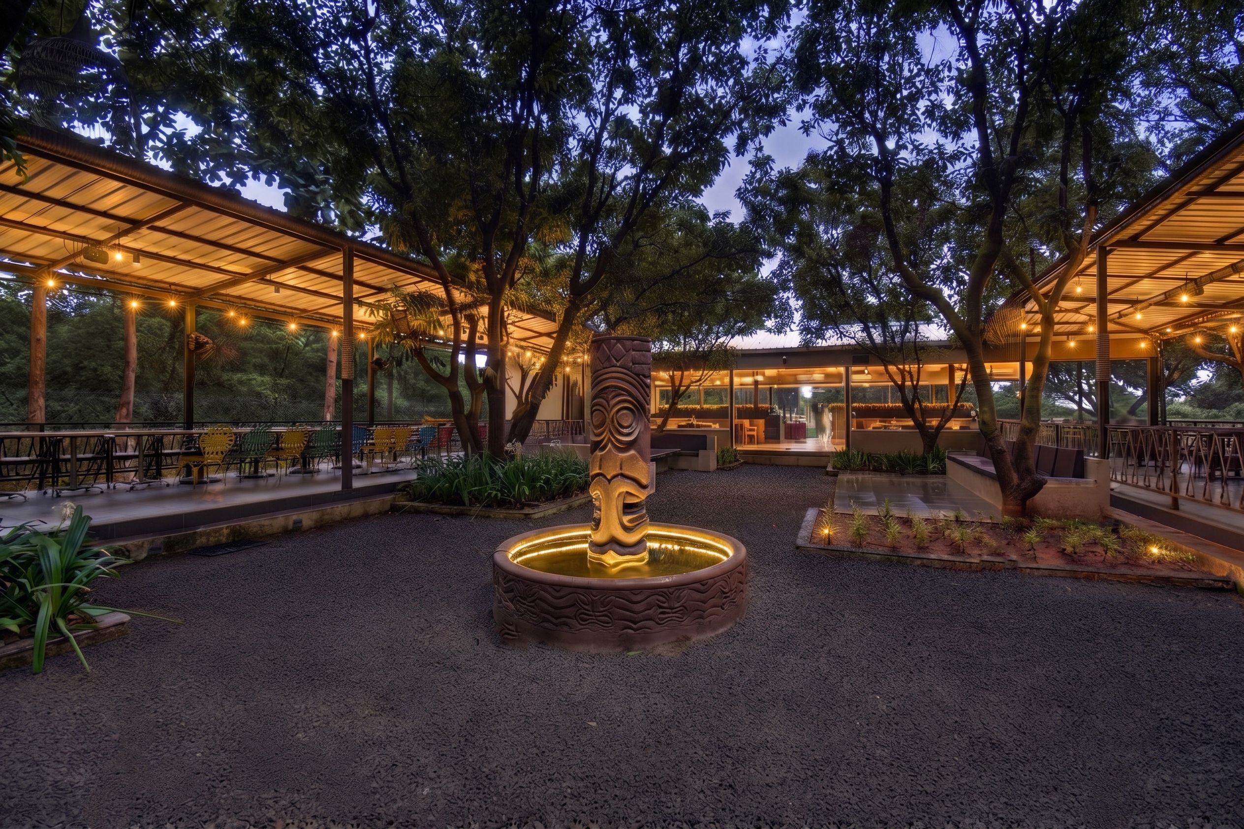 An outdoor seating area with tables and chairs, surrounded by trees and a central decorative feature.