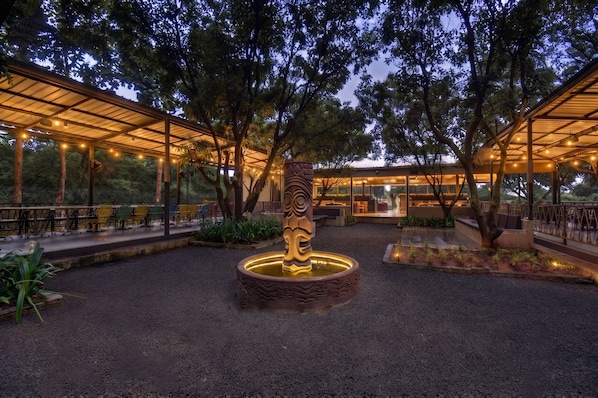 Un área de estar al aire libre con mesas y sillas, rodeada de árboles y un elemento decorativo central.