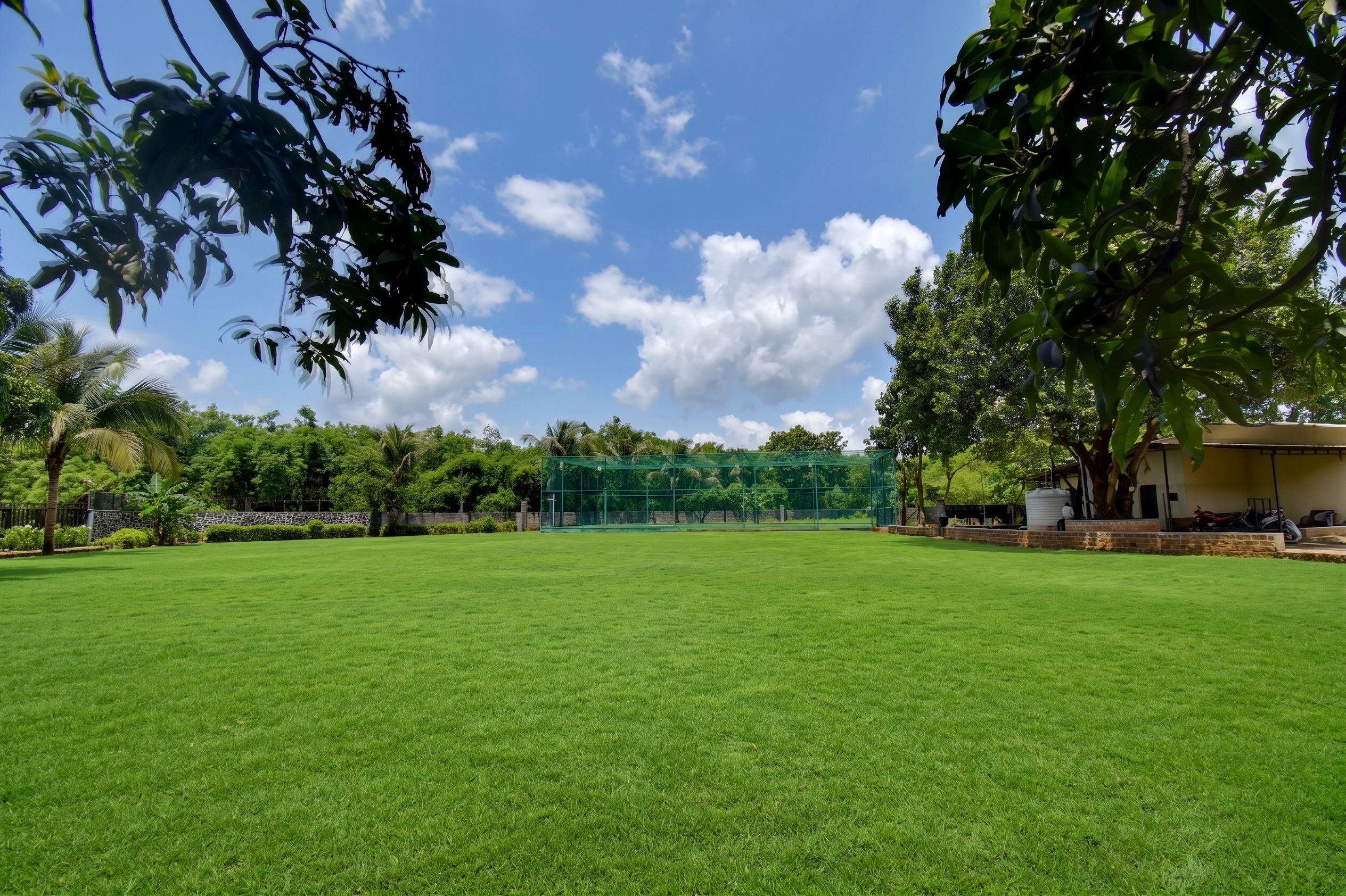 Um campo gramado bem cuidado, com uma área cercada por rede verde ao fundo, rodeado por árvores exuberantes e céu claro.