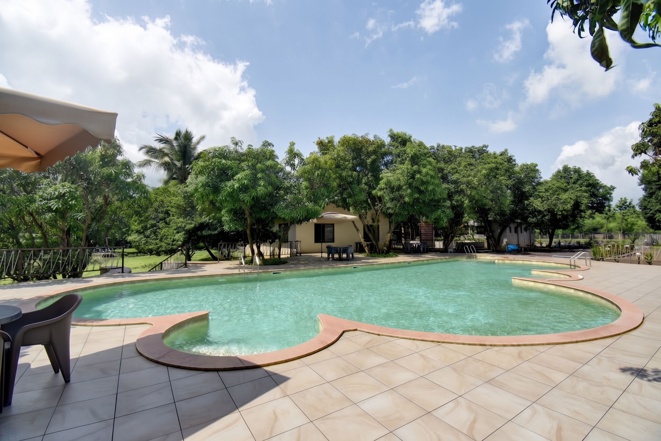 Una piscina con borde curvo, rodeada por un solárium con baldosas, mobiliario exterior y una zona de estar cubierta.