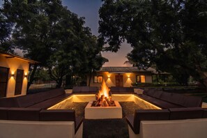 An outdoor seating area with a fire pit in the center, surrounded by sofas and a building with a thatched roof in the background. - Tiki Farms – Polynesian Boutique Resort, Karjat (Karjat)