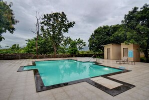 An outdoor swimming pool with a tiled deck, surrounded by trees and a small building with a door and windows. - Tiki Farms – Polynesian Boutique Resort, Karjat (Karjat)