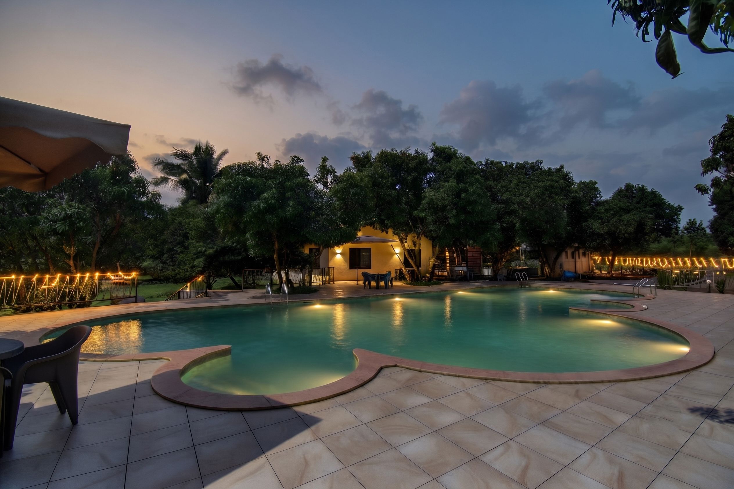 Una piscina con borde curvo, rodeada por un solárium con baldosas, y un pequeño edificio con un accesorio de iluminación al fondo.